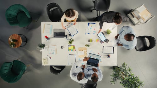 Vista cenital de los directivos revisando datos y propuestas durante la reunión corporativa. Un equipo diverso debate los objetivos, los presupuestos y los indicadores para la gestión de la empresa.